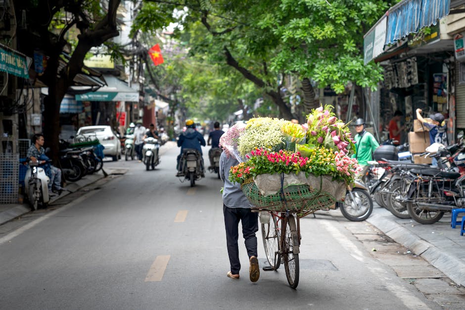 A street scene featuring a person pushing a bicycle loaded with a large, colorful floral arrangement. The bouquet includes pink roses, white hydrangeas, yellow lilies, and green foliage, arranged in a
