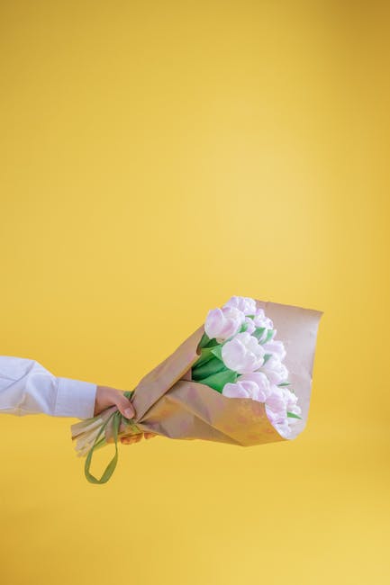 A person wearing a white shirt holds a bouquet of fresh pale pink tulips wrapped in light brown craft paper with a green ribbon, against a vibrant yellow background. The tulips have smooth, rounded pe