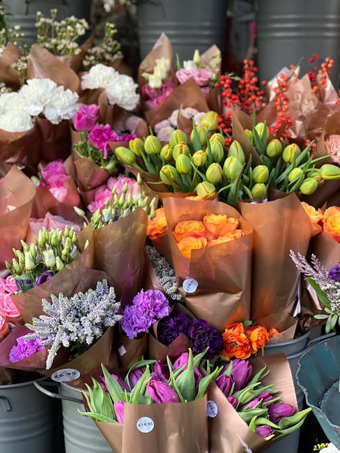 A vibrant display of fresh flowers arranged in brown tissue paper wraps, featuring various blooms including pink and purple tulips, orange roses, white and purple blossoms, and green buds. The bouquet