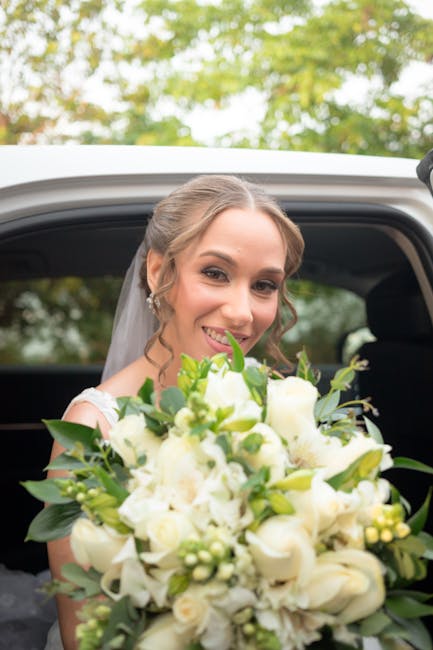 A bride with fair skin and brown hair styled in loose curls, smiling warmly while holding a large, lush bouquet of predominantly white flowers, including roses, lilies, and small filler blooms, accent
