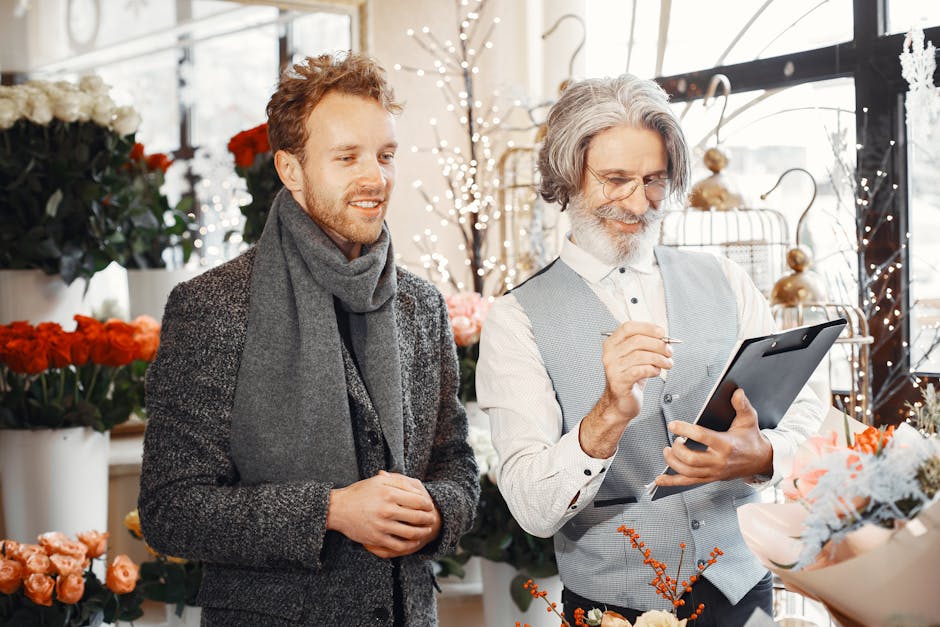 A professional florist with gray hair and a beard, wearing a light gray vest and white shirt, is holding a tablet and appears to be explaining something to a young man with reddish-brown hair and a be