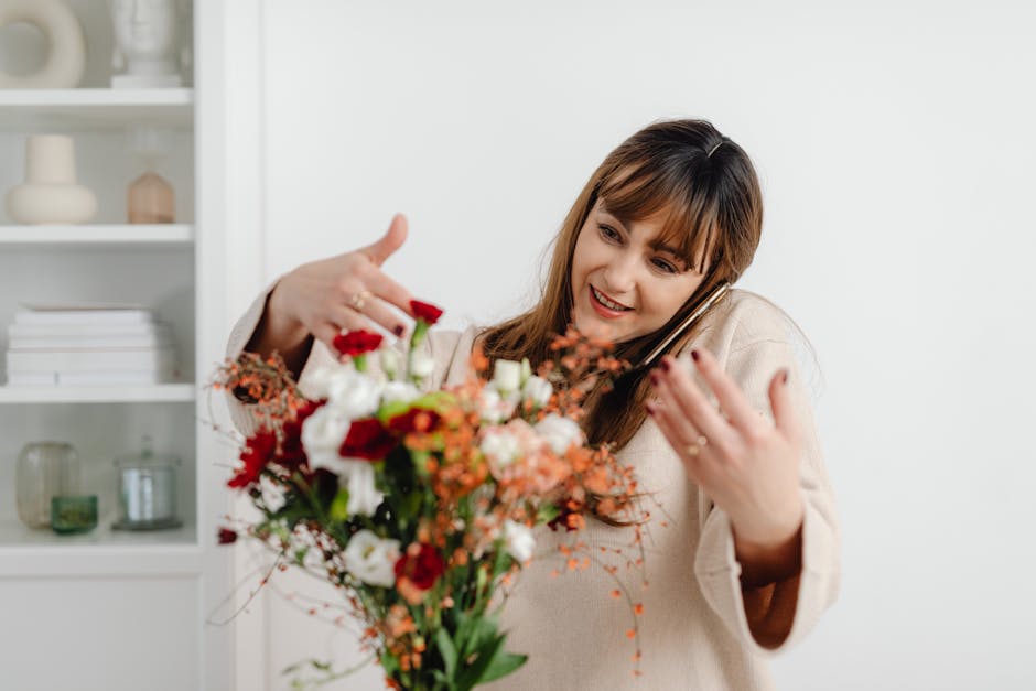 A woman with light brown hair and a beige sweater smiling happily while holding a vibrant floral bouquet close to her face. The bouquet features a mix of fresh flowers including red roses, white lisia