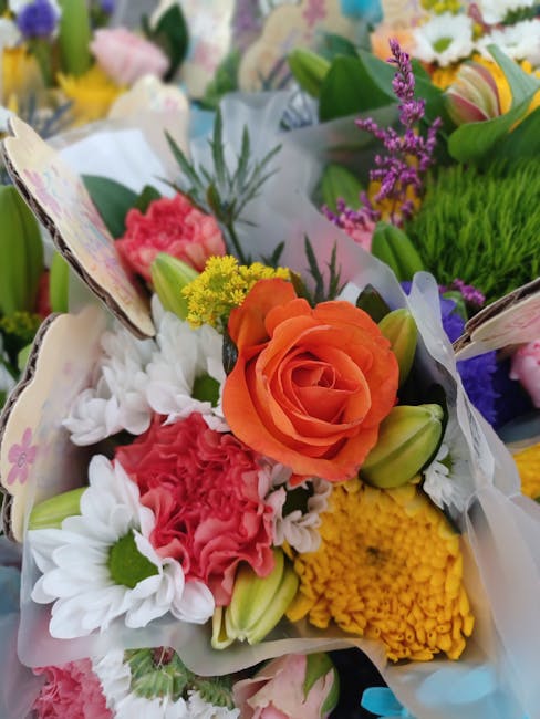 A vibrant floral bouquet featuring an orange rose as the focal point, surrounded by white daisies, pink carnations, yellow chrysanthemum, and purple lavender sprigs. The flowers are arranged with gree
