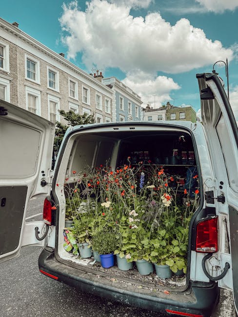 The open back of a white delivery van filled with potted plants and freshly cut flowers, including red poppies and white blooms, arranged in blue and gray pots. The flowers have vibrant colors and app