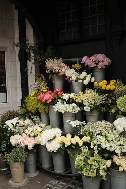 A variety of fresh floral arrangements displayed in metallic buckets on a dark outdoor wall, featuring predominantly pastel pink, white, and yellow blooms. The bouquet includes pink peonies, white ros